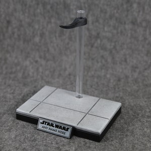 May include: A clear plastic display stand with a black base and a gray platform. The stand features a black support arm and a small rectangular sign that reads "STAR WARS ANY NAME HERE".