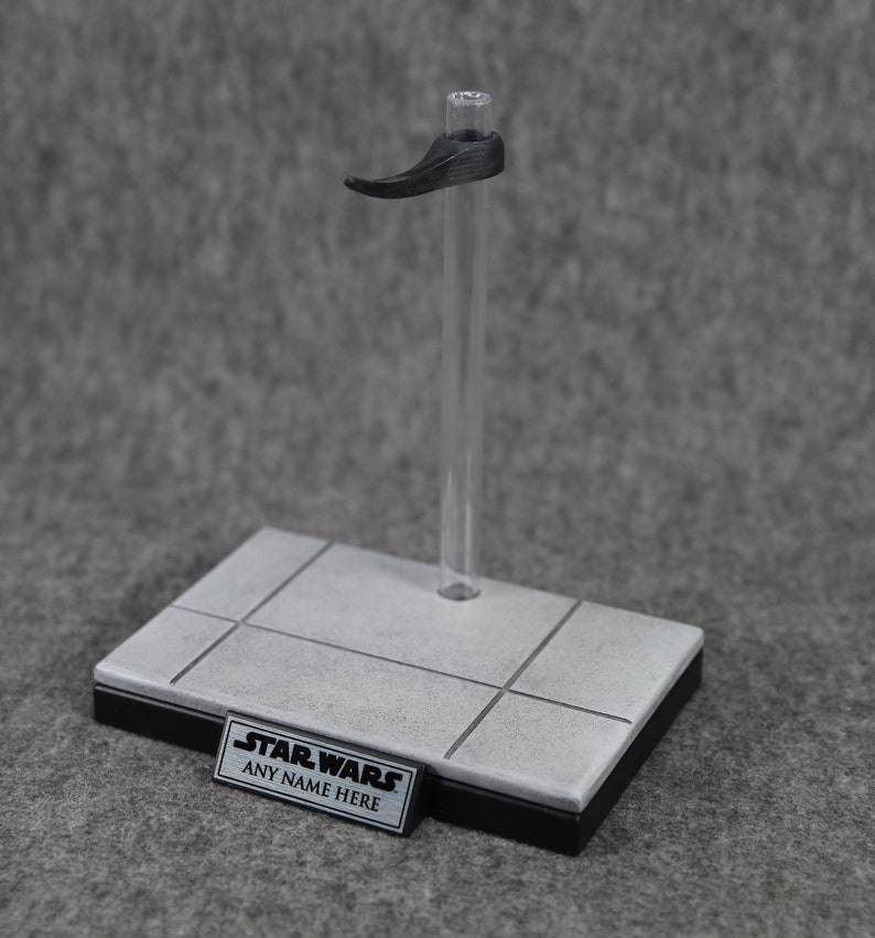 May include: A clear plastic display stand with a black base and a gray platform. The stand features a black support arm and a small rectangular sign that reads "STAR WARS ANY NAME HERE".