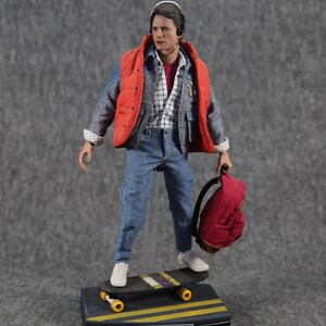K&ouml;nnte beinhalten: Eine Figur tr&auml;gt eine rote Weste, eine blaue Jeansjacke und eine blaue Jeans. Sie h&auml;lt einen roten Rucksack und steht auf einem Skateboard. Die Figur tr&auml;gt ein Headset und hat braunes Haar. Das Skateboard hat gelbe R&auml;der und ein schwarzes Deck. Die Figur steht auf einem schwarzen Sockel mit den Worten "Zur&uuml;ck in die Zukunft" und "Marty McFly" darauf.