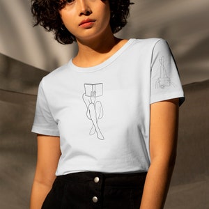 Op de afbeelding: Een lichtblauw t-shirt met een lijntekening van een persoon die met een boek op hun hoofd zit. Het shirt heeft een lijntekening van iemands benen op de rechtermouw.