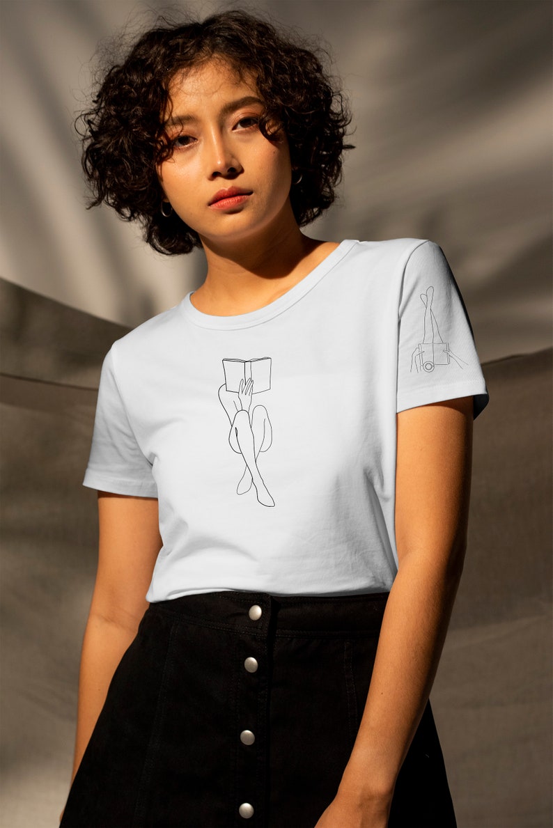 Op de afbeelding: Een lichtblauw t-shirt met een lijntekening van een persoon die met een boek op hun hoofd zit. Het shirt heeft een lijntekening van iemands benen op de rechtermouw.