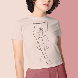 Op de afbeelding: Een wit T-shirt met een zwarte lijntekening van een persoon die met een boek op hun hoofd zit.