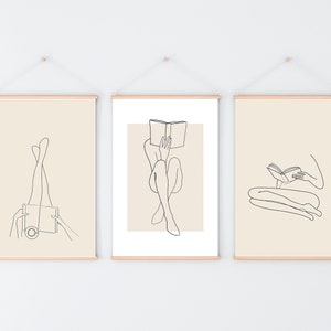 May include: Three minimalist line art prints featuring a person reading a book in different poses. The prints are framed with a light wood frame and hung on a white wall.