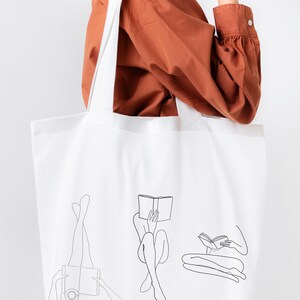Op de afbeelding: Een witte tote bag met een zwarte lijntekening van drie mensen die boeken lezen. De tas heeft een lange band en wordt gedragen door iemand die een bruin shirt draagt.