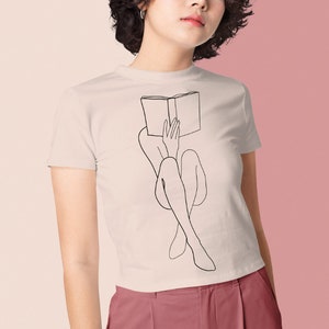 Op de afbeelding: Een wit T-shirt met een zwarte lijntekening van een persoon die met een boek op hun hoofd zit.