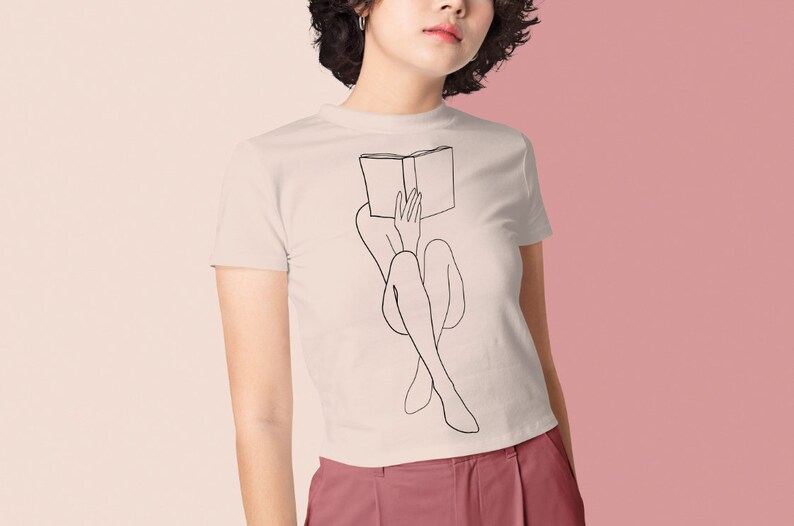 Op de afbeelding: Een wit T-shirt met een zwarte lijntekening van een persoon die met een boek op hun hoofd zit.