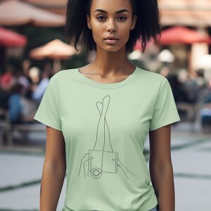 Op de afbeelding: Een lichtgroen T-shirt met een lijntekening van benen die uit een boek steken. Het boek heeft een cirkel op de voorkant.