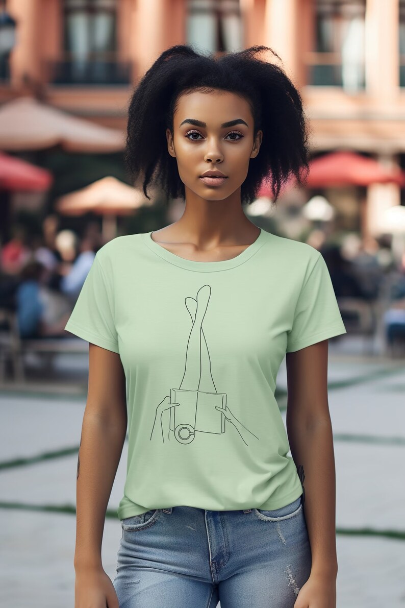 Op de afbeelding: Een lichtgroen T-shirt met een lijntekening van benen die uit een boek steken. Het boek heeft een cirkel op de voorkant.