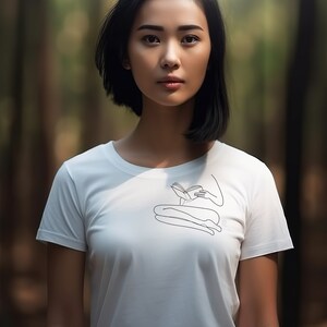 Op de afbeelding: Een vrouw met kort zwart haar draagt een wit T-shirt met een lijntekening van een persoon die zit en een boek leest. De vrouw staat in een bos.