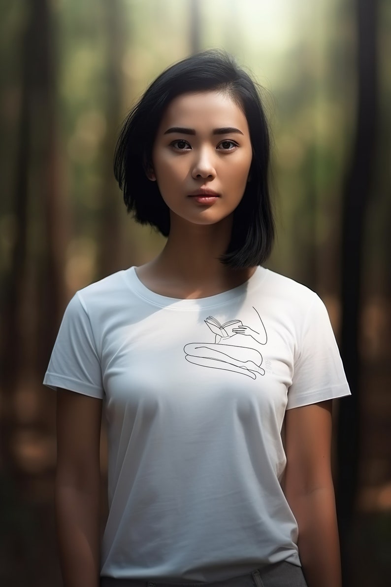 Op de afbeelding: Een vrouw met kort zwart haar draagt een wit T-shirt met een lijntekening van een persoon die zit en een boek leest. De vrouw staat in een bos.