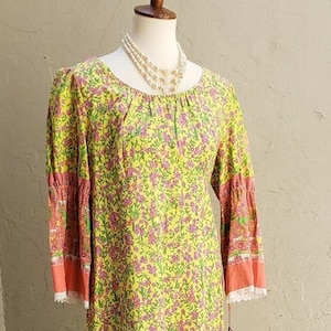 May include: A yellow floral print dress with long sleeves and a scoop neckline. The sleeves have pink and orange accents with white lace trim. A multi-strand pearl necklace is draped on the mannequin.