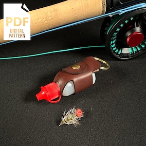 Könnte beinhalten: Ein brauner Lederflaschenhalter mit einem Messing-Druckknopfverschluss und einem Schlüsselring. Die Flasche ist klar mit einem roten Verschluss und mit Flüssigkeit gefüllt. Der Flaschenhalter ist an einer Angelrute befestigt.