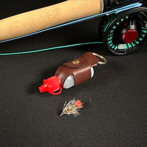 Könnte beinhalten: Ein brauner Lederhalter für einen Fliegenfisch-Linienkonditionierer mit roter Kappe und silbernem Schlüsselring. Der Halter ist an einer durchsichtigen Plastikflasche befestigt.