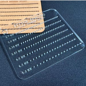 K&ouml;nnte beinhalten: Ein transparentes Acryl-Stichabstandswerkzeug mit Messungen in Millimetern. Das Werkzeug hat eine Reihe von gleichm&auml;&szlig;ig verteilten L&ouml;chern und den Text "STITCH SPACING" und "SPACE BETWEEN HOLES". Die Messungen reichen von 2,7 mm bis 5,0 mm.