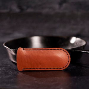 Könnte beinhalten: Ein brauner Topfhandschuh aus Leder mit schwarzen Nähten. Der Topfhandschuh liegt vor einer schwarzen Gusseisenpfanne.