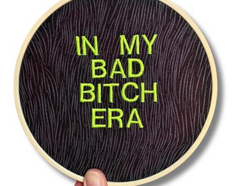 Puede incluir: Bastidor de bordado redondo con el texto "IN MY BAD BITCH ERA" en hilo verde neón. La tela tiene un fondo negro con un sutil patrón ondulado. El bastidor de madera está sostenido por una mano.