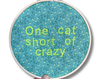 Puede incluir: Un bastidor de bordado redondo con un fondo de tela turquesa con un estampado floral. El texto "One cat short of crazy" está bordado en hilo amarillo brillante. El bastidor es sostenido por una mano.