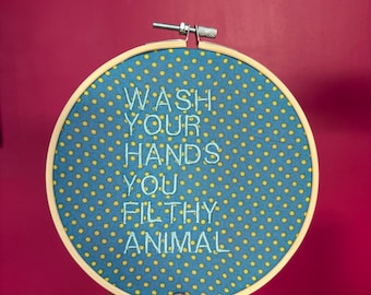 Puede incluir: Aro bordado con el texto "WASH YOUR HANDS YOU FILTHY ANIMAL" en hilo azul claro. La tela es azul con lunares amarillos. El aro es de madera y tiene un mecanismo de apriete metálico.
