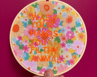 Puede incluir: Un bastidor de bordado redondo con un fondo de tela floral. El texto "WASH YOUR HANDS YOU FILTHY ANIMAL" está bordado con hilo naranja. El bastidor se sostiene sobre un fondo magenta.