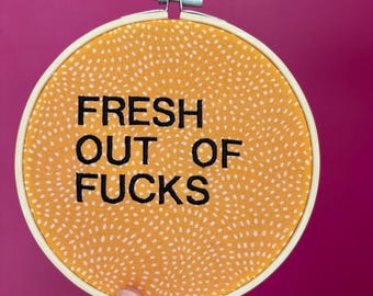 Puede incluir: Un bastidor de bordado redondo con el texto "FRESH OUT OF FUCKS" en letras negras. La tela dentro del bastidor es naranja con pequeños puntos blancos. El bastidor se sostiene sobre un fondo magenta.