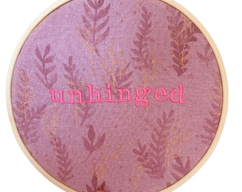 Puede incluir: Un bastidor de bordado redondo con un fondo de tela rosa con un patrón de hojas y la palabra "unhinged" bordada en hilo rosa. El bastidor de madera está sostenido por una mano.
