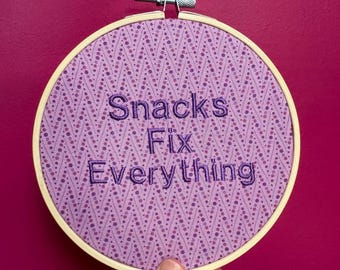 Puede incluir: Un bastidor redondo con una tela morada con un patrón de puntos y zigzags. El texto "Snacks Fix Everything" está bordado con hilo morado. El bastidor se sostiene sobre un fondo magenta.