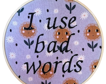 Puede incluir: Un bastidor de bordado redondo con tela lila claro con un estampado floral y el texto "I use bad words" en negro. El diseño incluye girasoles sonrientes y flores blancas parecidas a margaritas.