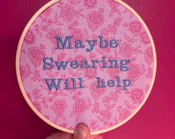 Puede incluir: Un bastidor de bordado redondo con tela estampada floral rosa. El texto "Maybe Swearing Will Help" está bordado en hilo azul. El bastidor se sostiene sobre un fondo rosa.