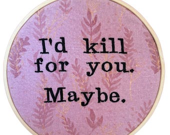 Puede incluir: Aro de bordado redondo con un fondo de tela rosa con un patrón de hojas y el texto "I'd kill for you. Maybe." El texto está en hilo negro. El aro es de madera y tiene un mecanismo de apriete metálico.
