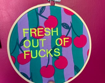 Puede incluir: Aro de bordado redondo con un diseño de cerezas y el texto "FRESH OUT OF FUCKS" en verde neón. El fondo tiene rayas verticales en verde azulado y morado. Las cerezas son de color rosa brillante.
