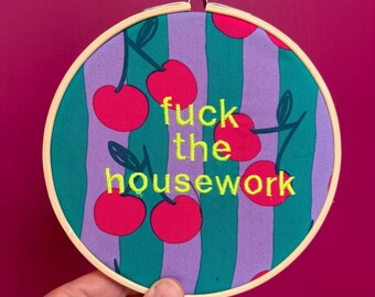 Puede incluir: Un bastidor redondo con un diseño de tela con cerezas y rayas verticales verdes y moradas. El texto "fuck the housework" está bordado con hilo amarillo.