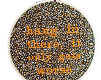 Puede incluir: Bastidor de bordado redondo con tela floral en negro, amarillo y blanco. El texto "hang in there, it only gets worse" está bordado en hilo naranja. El bastidor es sostenido por una mano.