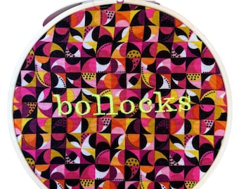 Puede incluir: Un bastidor de bordado con un vibrante patrón geométrico en rosa, naranja, amarillo y negro. La palabra "bollocks" está bordada con hilo verde. El bastidor es sostenido por una mano, sobre un fondo blanco.