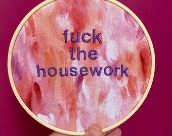 Puede incluir: Bastidor de bordado redondo con un diseño de tela en tonos rosa, naranja y blanco. La frase "fuck the housework" está bordada con hilo morado. El bastidor se sostiene sobre un fondo magenta.