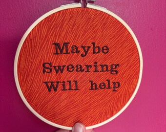 Puede incluir: Un bastidor de bordado con tela naranja y el texto "Maybe Swearing Will help" en negro. La tela tiene un patrón texturizado. El bastidor se sostiene sobre un fondo rosa.