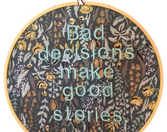 Puede incluir: Un bastidor redondo con la frase "Bad decisions make good stories" bordada en hilo azul claro. El fondo de tela es de color verde azulado oscuro con un estampado floral en tonos dorados, grises y morados. El bastidor de madera está sujeto por una mano.