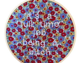 Puede incluir: Aro de bordado redondo con fondo de tela floral. La tela presenta flores rojas, azules y amarillas. El texto bordado en blanco dice "It's a full time job being a bitch."