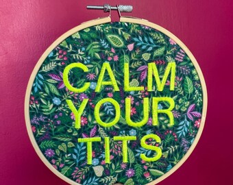 Puede incluir: Un aro de bordado redondo con el texto "CALM YOUR TITS" en hilo verde brillante. El fondo de tela presenta un patrón floral verde oscuro con acentos azules, rosas y morados. El aro es sostenido por una mano sobre un fondo magenta.