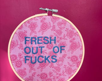 Puede incluir: Un bastidor de bordado redondo con tela floral rosa y el texto bordado en azul "FRESH OUT OF FUCKS". El bastidor de madera se encuentra sobre un fondo rosa.
