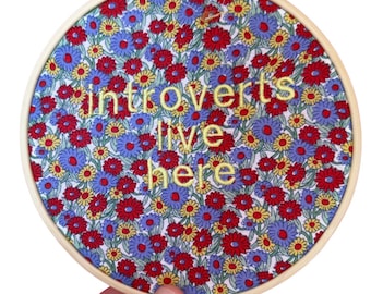 Puede incluir: Un bastidor de bordado redondo con un diseño de tela floral con flores rojas, azules y amarillas. El texto "introverts live here" está bordado con hilo amarillo. El bastidor es sostenido por una mano.