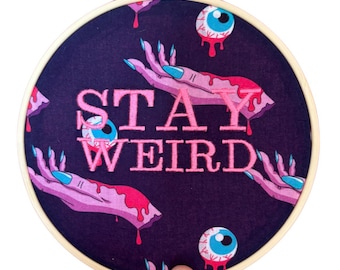 Puede incluir: Un bastidor redondo con tela morada oscura con la frase rosa "STAY WEIRD". El diseño incluye manos rosas con uñas azules, sangre roja goteando y globos oculares azules. El bastidor está sostenido por una mano.