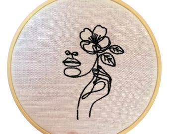 Puede incluir: Diseño bordado en negro sobre tela blanca dentro de un bastidor de madera. El diseño presenta un dibujo lineal minimalista de un rostro con una flor y una mano. El bastidor es sostenido por una mano.