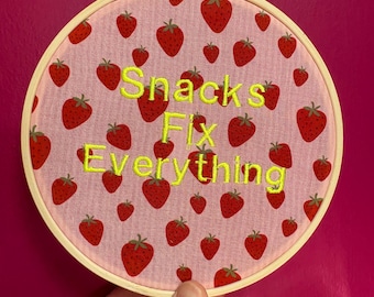 Puede incluir: Aro de bordado redondo con un fondo de tela rosa con un patrón de fresas rojas. El texto "Snacks Fix Everything" está bordado en hilo amarillo. El aro de madera está sostenido por una mano sobre un fondo rosa.