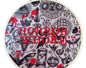 Puede incluir: Aro de bordado redondo con un diseño de tela con símbolos ocultos en negro y rojo. Las palabras "HORROR WHORE" están bordadas en hilo rojo. El diseño incluye un ojo, un reloj de arena y otros elementos místicos.