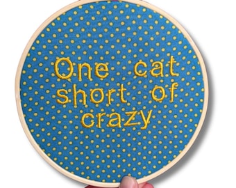 Puede incluir: Aro de bordado redondo con un fondo de tela azul con lunares amarillos. El texto "One cat short of crazy" está bordado con hilo amarillo. El aro es sostenido por una mano.