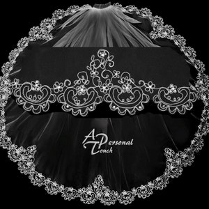 May include: A white bridal veil with a delicate lace trim and floral embroidery. The lace is adorned with small, sparkling rhinestones. The veil is a single layer and has a scalloped edge.