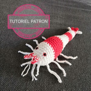 K&ouml;nnte beinhalten: Geh&auml;kelte Garnele mit rot-wei&szlig;en Streifen, wei&szlig;en F&uuml;hlern und Beinen. Die Garnele hat kleine rote Augen und liegt auf einer dunkelgrauen Oberfl&auml;che. Der Text "TUTORIEL PATRON" befindet sich in einem rosa Kreis im Hintergrund.