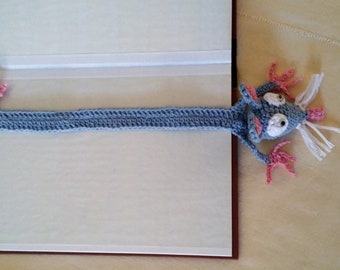 May include: A handmade crochet bookmark in the shape of a cartoon rat. The rat is blue with pink accents on its ears, feet, and tail. It has white whiskers and large, expressive eyes. The bookmark is placed in a book.