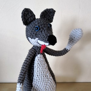 Peut inclure: Un jouet en crochet en forme de loup gris et blanc avec des yeux bleus et une langue rouge qui sort. Le loup est debout sur une surface en bois et a une patte levée en l'air.