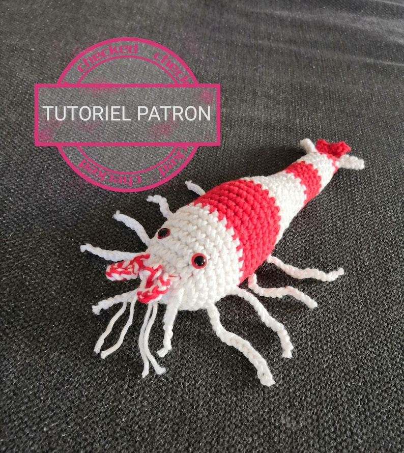 K&ouml;nnte beinhalten: Geh&auml;kelte Garnele mit rot-wei&szlig;en Streifen, wei&szlig;en F&uuml;hlern und Beinen. Die Garnele hat kleine rote Augen und liegt auf einer dunkelgrauen Oberfl&auml;che. Der Text "TUTORIEL PATRON" befindet sich in einem rosa Kreis im Hintergrund.